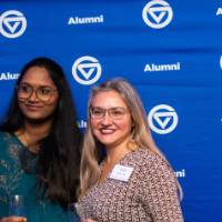 Two grads smile and hold glasses in front of backdrop